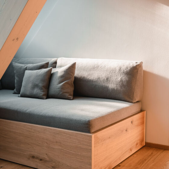 Gemütliche Sitzbank mit grauen Kissen unter Dachschräge aus Holz.