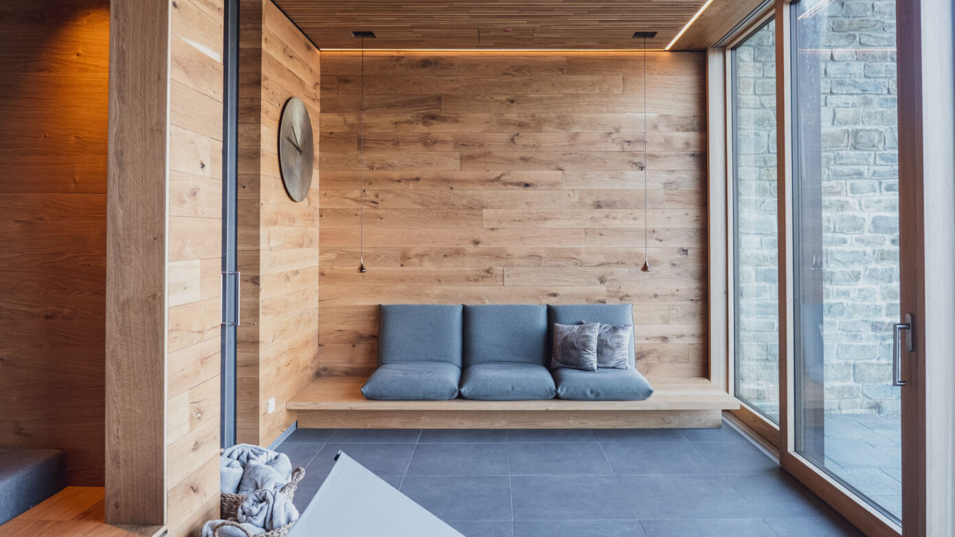 Moderner Sauna und Wellness Bereich mit Holzverkleidung, grauem drei-Sitzer Sofa und großen Fenstern. Links ist die Tür zur Sauna, rechts die Tür zur Außenterrasse. Körber mit Handtücher liegen am Boden.