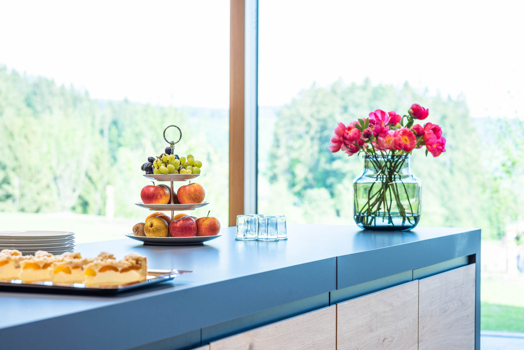 Frisches Obst und Blumen auf einer blauen Küchentheke vor Fenster mit Blick ins Grüne.