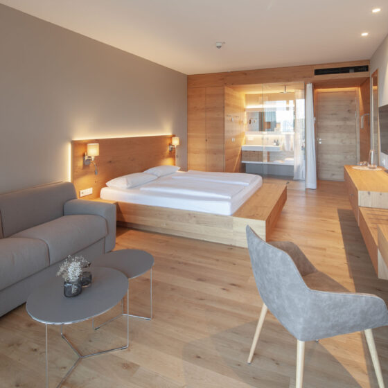 Moderne Hotelzimmer mit Doppelbett, Sofa, Schreibtisch und Blick auf Badezimmer mit Glaswand.