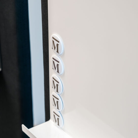 Mehrere weiße Magnete mit schwarzen "M"-Zeichen an einer weißen Oberfläche.