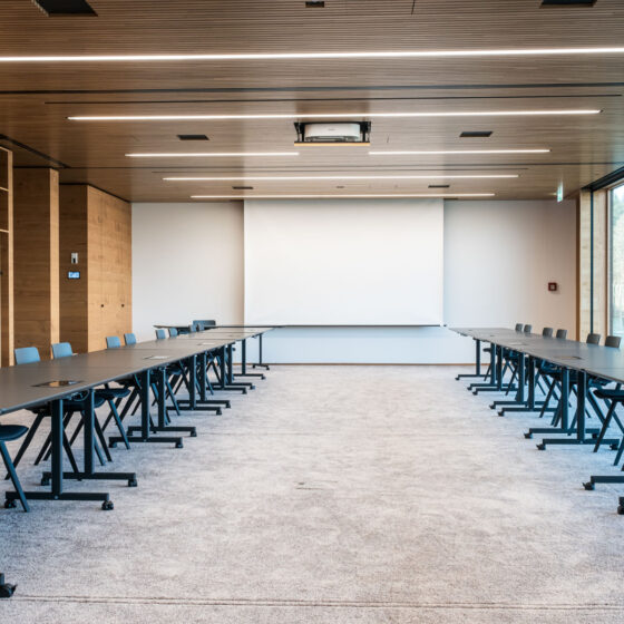 Moderner Konferenzraum mit Tischen, Stühlen und großem Whiteboard, viel Tageslicht durch Fenster.