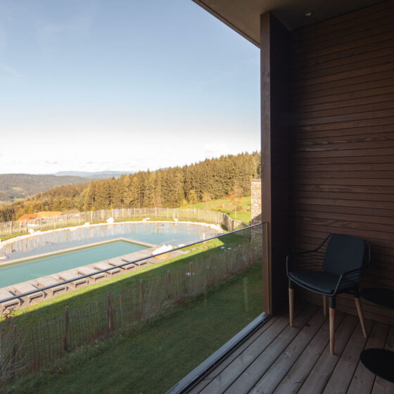 Blick von Balkon auf Pool und grüne Hügel mit Wald im Hintergrund, sonniger Tag.