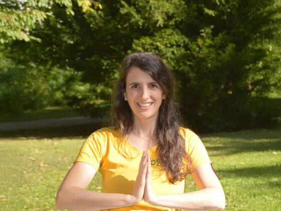 Lächelnde Frau in Yoga-Posen im Park bei Sonnenschein, umgeben von grünen Bäumen.