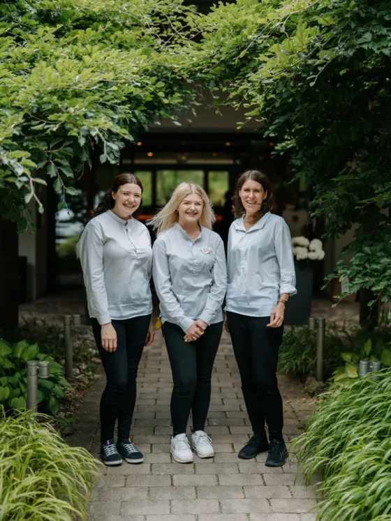 Drei lachende Frauen in weißen Hemden stehen auf einem Gartenweg unter grünen Bäumen.