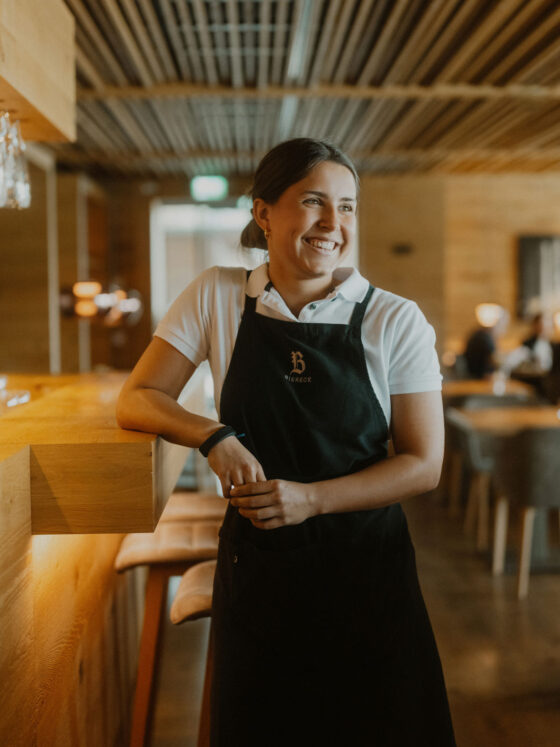 Lächelnde Kellnerin in einem Holzrestaurant, trägt schwarze Schürze und weißes Polo.