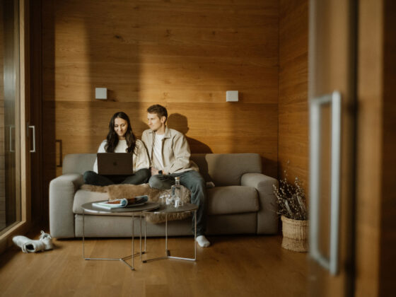 Zwei junge Menschen sitzen auf einem Sofa mit Laptop in einem gemütlichen, holzverzierten Wohnzimmer.