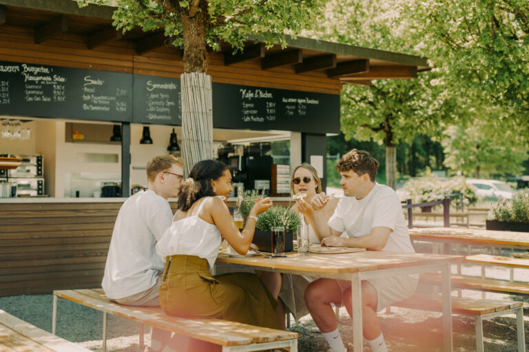 Freunde essen und trinken bei schönem Wetter im Außenbereich eines Restaurants unter Bäumen.